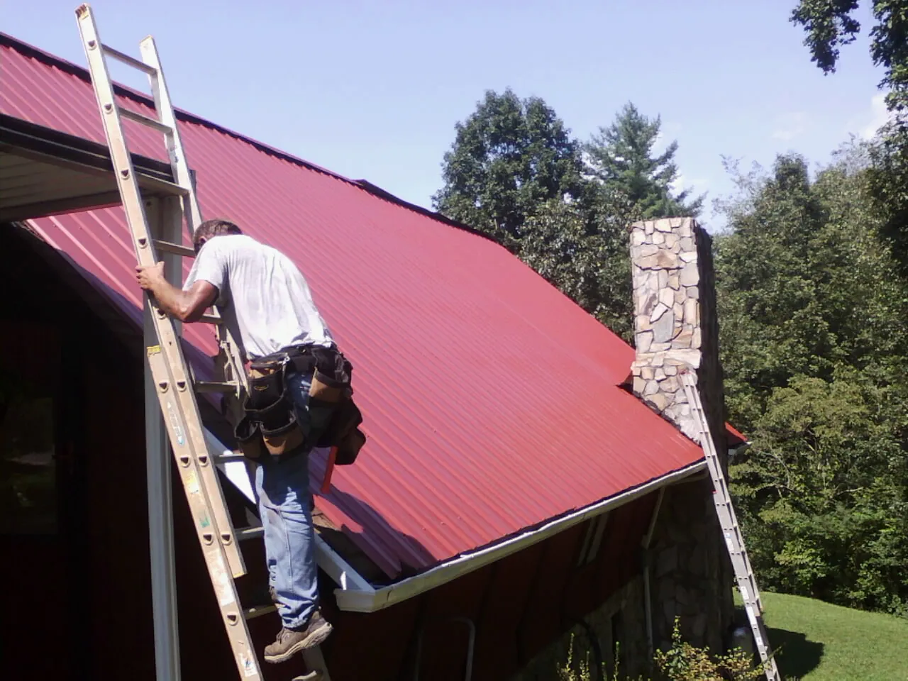 Roofer on a ladder beside a red metal roof and stone chimney — Soffit & Fascia Repair by Roofing Brothers in Cable